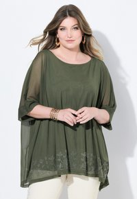 Blusa trasparente verde oliva con maniche larghe e ricami floreali lungo l'orlo; il design a strati offre una vestibilità rilassata sopra dei pantaloni bianchi.