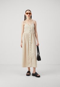 Beige len maxi ruha vékony pántokkal, gombos elülső résszel és ráncolt derékkal. Fekete platform szandállal és egy kis fekete táskával párosítva.