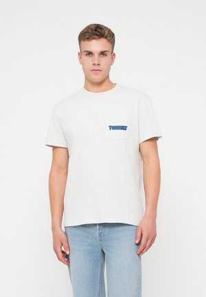 Noor mees kannab heledat halli T-särki, millel on rindade taskul väike sinine "Tommy Jeans" logo, ja hele sinine teksapüksid, seistes valge tausta ees.