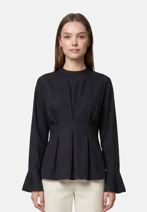 Vrouw draagt een zwarte blouse met lange mouwen en uitlopende manchetten en beige broek, staand tegen een effen lichte achtergrond.
