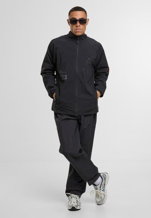 Homme portant une veste noire zippée, un pantalon noir, des chaussettes blanches et des baskets grises, debout avec les mains dans les poches et les jambes croisées, portant des lunettes de soleil.