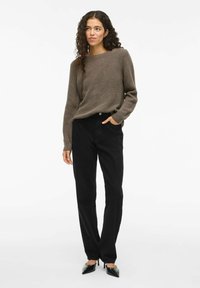 Femme aux cheveux bouclés portant un pull en tricot marron, un pantalon noir taille haute et des escarpins noirs à bout pointu, se tenant avec une main dans la poche.