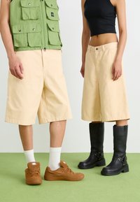 Pantaloni corti larghi beige, gilet utility verde con tasche, sneakers marroni e stivali neri fino al ginocchio su un pavimento verde.