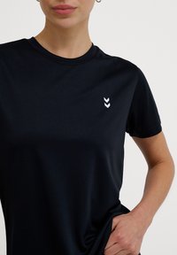 Kvinde iført en ensfarvet sort T-shirt med korte ærmer med to hvide pil-chevron-logoer på venstre bryst, hånd hvilende på taljen.