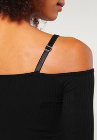 Top nero a costine off-the-shoulder con spalline sottili regolabili e un leggero lucido. Texture del tessuto visibile con dettagli a costine orizzontali.