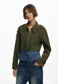 Olijfgroen cropped jack met een rits aan de voorkant en schouderepauletten, gecombineerd met blauwe denim high-waisted broeken met voorzakken.