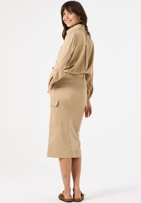 Beige Bluse und knielanger Rock aus weichem Stoff. Der Rock hat seitliche Taschen und wird mit flachen Sandalen kombiniert.