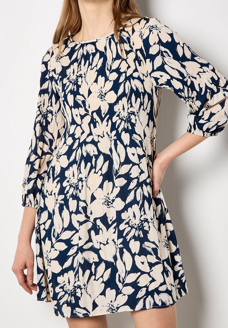 Femme portant une robe bleu marine avec un imprimé floral beige et des manches bouffantes trois-quarts, debout la main sur la hanche.