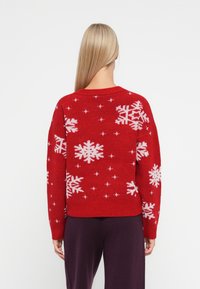 Pull en maille rouge avec des motifs de flocons de neige et d'étoiles blancs, col rond, ourlet côtelé et manches longues. Porté sur un pantalon violet foncé.