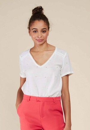 Blouse blanche à col V avec des manches courtes retroussées, ornée de petits motifs triangulaires dorés, associée à un pantalon ajusté rose vif.