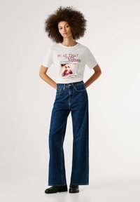 Bela grafična majica s tekstom in sliko, kombinirana z visokim pasom in širokimi modrimi kavbojkami. Obutev dopolnjujejo črni čevlji.