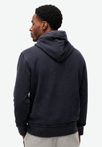 Granatowy hoodie wykonany z mieszanki bawełny, z kapturem na sznurki, ściągaczami przy rękawach i luźnym krojem, widoczny od tyłu.