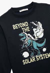 Czarna bluza z grafiką przedstawiającą dinozaura w skafandrze kosmicznym oraz astronautę, z napisem "POZA UKŁADEM SŁONECZNYM" w kolorze żółtym.