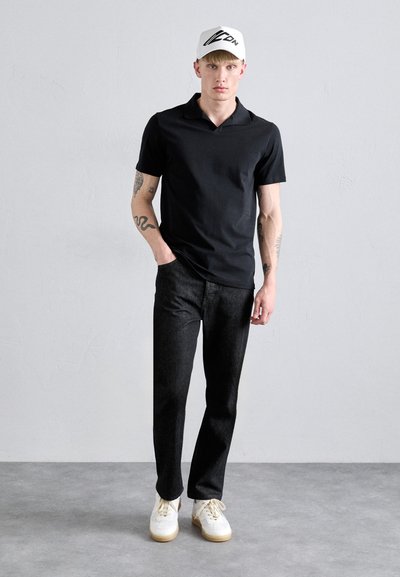 Polo shirt nera abbinata a jeans scuri e sneakers bianche. Il modello indossa un cappellino bianco con logo nero. Design semplice e casual, silhouette aderente.