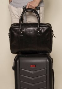 Sort læderhandtaske med dobbelte håndtag hviler på en sort hårdskalskuffert med struktureret overflade og en rød logoaccent.
