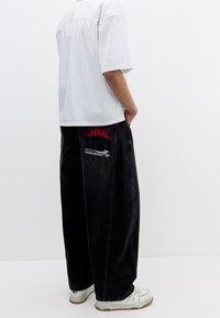 Bílé pruhované tričko s krátkým rukávem, volně střižené černé džíny s červeným grafickým a ručně malovaným motivem na zadní kapse, doplněné bílými teniskami.