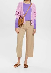 Ljust rosa stickad cardigan över en lila topp, kombinerad med beigefärgade vida byxor och bruna sandaler. Axelväska i brunt läder.