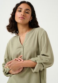 Bluză verde măsline, cu nasturi, cu decolteu în V, mâneci lungi suflecate la manșete și un material texturat. Design simplu cu o taie lejeră.