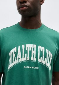 Άνδρας με πράσινο T-shirt που φέρει μεγάλο λευκό κείμενο "HEALTH CLUB" και μικρότερα γράμματα "BJÖRN BORG" κάτω από αυτό, με απλό φωτεινό φόντο.