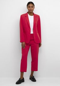 Blazer fucsia con colletto a punta, chiusura con un solo bottone; pantaloni cropped coordinati. T-shirt bianca sotto. Mocassini neri.