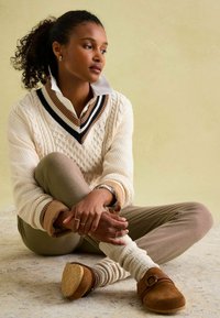 Cremefarbener gestrickter Pullover mit V-Ausschnitt und gestreiften Akzenten, beiger gemusterter Hose, hellen Stricksocken und braunen Clogs mit Schnallendetail.