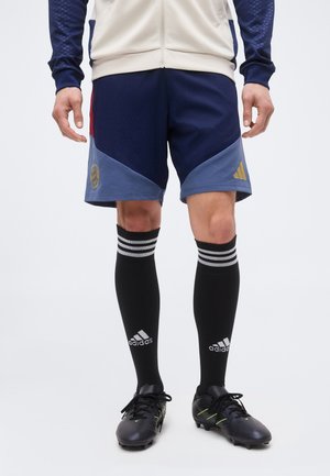 Homme portant un short de sport bleu marine et bleu avec le logo de l'équipe, des chaussettes noires montantes Adidas et des crampons, debout sur fond blanc.