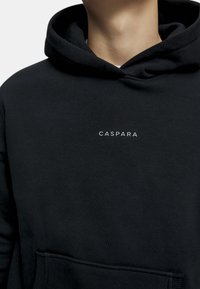 Musta huppari, joka on valmistettu pehmeästä kankaasta, varustettuna kengurupussilla ja rinnassa valkoisella "CASPARA"-logolla. Yksinkertainen muotoilu ja rento istuvuus.