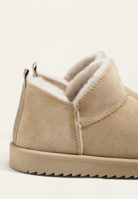 Beige mockasiner med en mjuk, krämfärgad pälsfoder. Har en rundad form, sydda detaljer och en texturerad gummisula med ett zigzag-mönster.