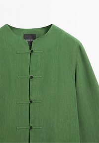 Zelená košile s dlouhým rukávem, bez límce a se čtyřmi černými zapínacími knoflíky na přední straně.