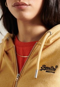 Sudadera con cremallera amarilla de tejido texturizado, con cordones, herrajes plateados y el logo "Superdry" bordado en negro, superpuesta sobre una camiseta roja.