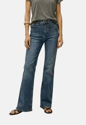 Mujer con vaqueros acampanados de tiro alto y lavado medio, camiseta gris, sandalias negras y una pulsera beige en la muñeca izquierda sobre un fondo liso.