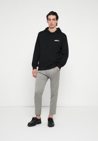 Felpa nera con cappuccio e tasca frontale, con logo colorato sul petto, abbinata a pantaloni slim-fit grigio chiaro e scarpe nere.