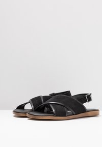 Black suede sandals feature crossed straps with shiny accents and a buckle. They have a flat sole and a textured brown heel.