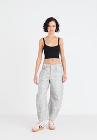Černý crop top v kombinaci s volnými kalhotami v světlé šedé barvě se vzorem. Model má na sobě růžové tenisky s černými pruhy. Jednoduché, minimalistické pozadí.