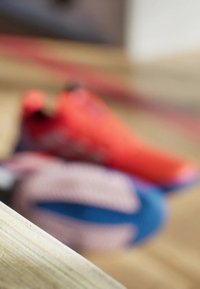 Czerwone buty sportowe z czarnymi akcentami ułożone na teksturowanej drewnianej powierzchni, rozmyte tło z niebieskimi i wzorzystymi butami.