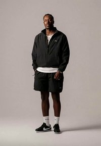 Casaco preto com fecho éclair sobre uma camisa branca, combinado com calções pretos. Usado com sapatilhas pretas e brancas e meias brancas de cano curto. Design simples.