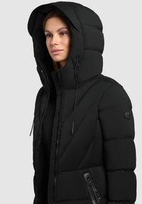 Žena v černé zateplené zimní bundě s velkou kapucí a kapsou na zip, pohledu vlevo na šedém pozadí.