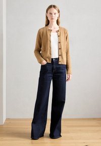 Cardigan marrone testurizzato con colletto tondo e tasche frontali, abbinato a jeans a gamba larga blu scuro a vita alta. Sfondo in legno naturale.