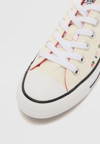 Krém színű vászon sneaker fehér fűzőkkel, piros varrással, fekete gumitalppal és hímzett eper részletekkel az oldalon.
