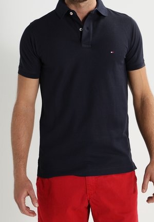 Man draagt een marineblauw polohemd met korte mouwen en een klein logo op de borst en felrode korte broek, staat met ontspannen armen.