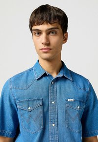 Blauwe denim overhemd met korte mouwen, voorzien van drukknoopsluiting, twee borstzakken met schuine kleppen en contrasterend stiksel. Wrangler-logo weergegeven.