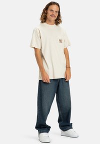 Beige bavlněné tričko s náprsní kapsou a hnědým logem, spárované s volně střiženými modrými džínami a bílými teniskami.