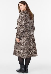 Vrouw in een knie-lange jurk met luipaardprint, zwarte maillots en zwarte enkellaarzen, staand en over haar schouder kijkend.