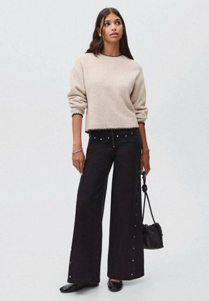 Pull beige avec bord noir, associé à un pantalon noir à jambes larges avec des boutons sur le côté. Le modèle tient un petit sac noir.