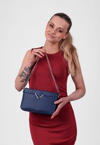 Tmavě modrá crossbody taška s řetízkem a stříbrným V-itém akcentem. Vyrobena z hladkého materiálu, s minimalistickým designem.