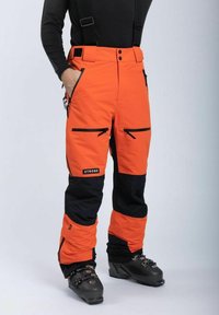 Oranssi ja mustat hiihtohousut säädettävillä olkaimilla, kahdella vetoketjullisella taskulla ja logopatchilla; teksturoitu kangas kestävyyden ja lämmön vuoksi.