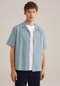Camicia a maniche corte, con bottoni, di colore azzurro chiaro con strisce bianche verticali, dotata di colletto e taschino sul petto. Indossata sopra una maglietta bianca.