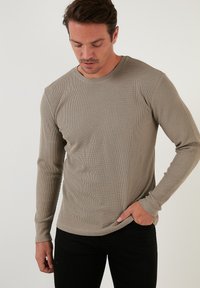 Beige long-sleeve thermal shirt with a textured ribbed pattern, round neckline, and fitted sleeves, paired with black trousers.