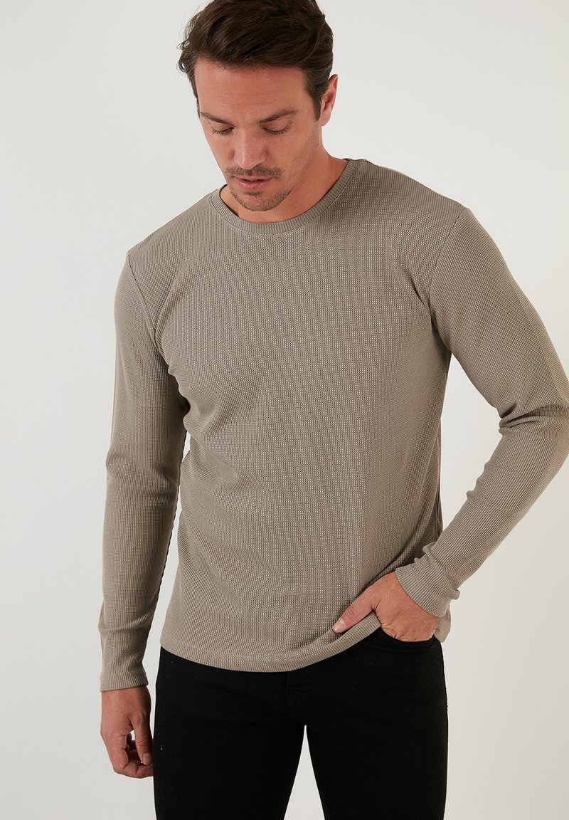 Beige long-sleeve thermal shirt with a textured ribbed pattern, round neckline, and fitted sleeves, paired with black trousers.