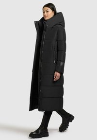 Schwarze Daunenjacke mit hohem Kragen, langem, gestepptem Design, Frontreißverschluss und seitlichen Taschen. Getragen mit schwarzen kniehohen Stiefeln.